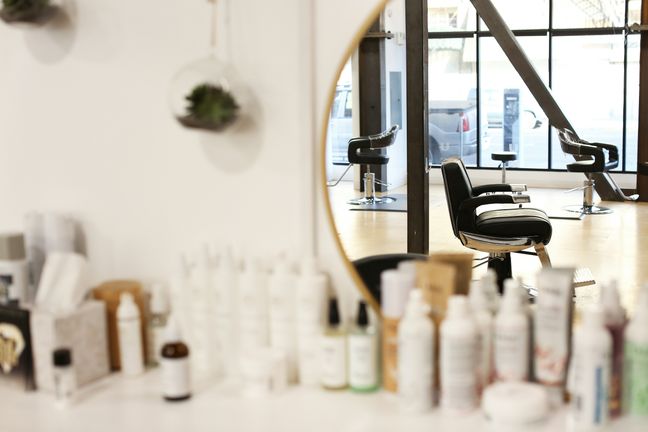 An empty barber chair reflected in a round mirror with a brass frame, hair products on the shelf in front